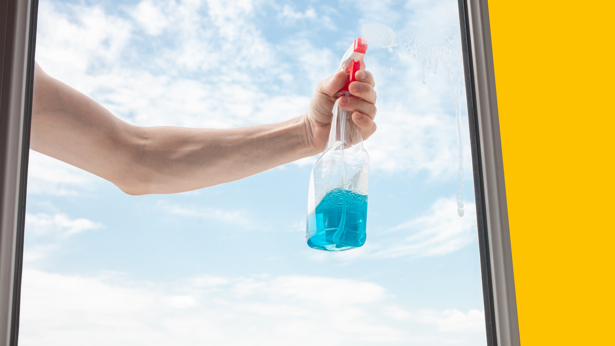 A man washes a window from the outside. The man sprays a blue cleaner