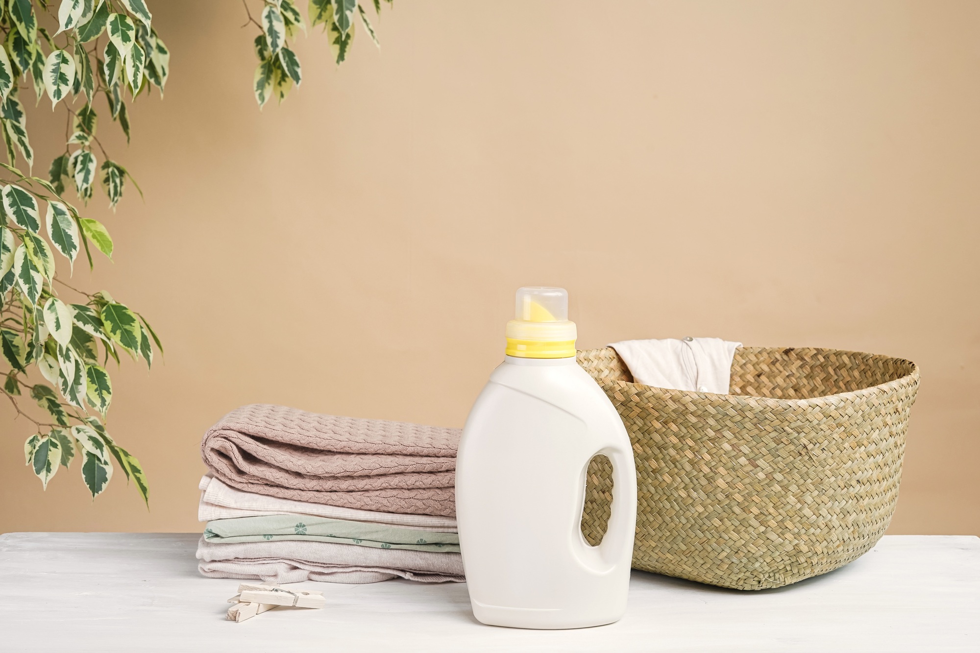 Laundry basket, stack of folded clothes, bottle of washing gel or fabric softener on a beige backgro